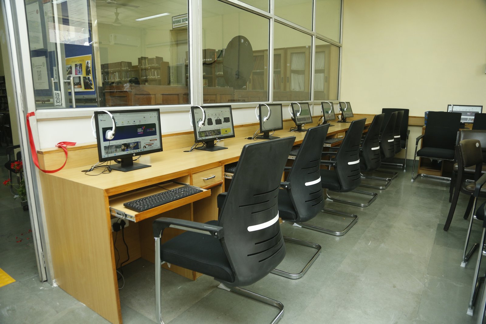A computer lab with a row of desktop computers placed on a long wooden desk. Each computer has a headset and a chair positioned in front of it.